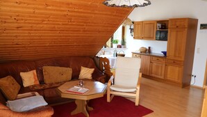 A cozy living area with a wooden ceiling, a leather sofa, a wooden table, and a kitchen with wooden cabinets and a microwave. - Ferienwohnung Däumler (Breitenbrunn/Erzgeb.)