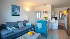A modern living room with a grey sofa, a blue armchair, and a coffee table with fruits and glasses. A kitchen area is visible in the background with a bar and a dining table. - Seetempel 34 Scharbeutz (Scharbeutz)
