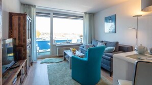 A modern living room with a sofa, armchair, and coffee table. A large window offers a view of a body of water and buildings. - Seetempel 34 Scharbeutz (Scharbeutz)