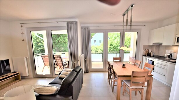 Un salon moderne comprenant un canapé, une table à manger et des chaises, de grandes portes-fenêtres coulissantes donnant sur un balcon, et un coin cuisine.
