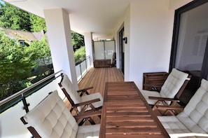 A balcony with a wooden table and four cushioned chairs, overlooking a garden and a building.