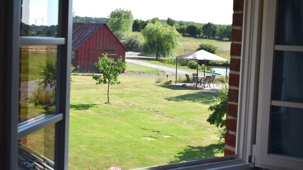 Une vue par la fenêtre d’une grange rouge, avec un petit arbre et un espace extérieur abrité, équipé de chaises et d’une table.