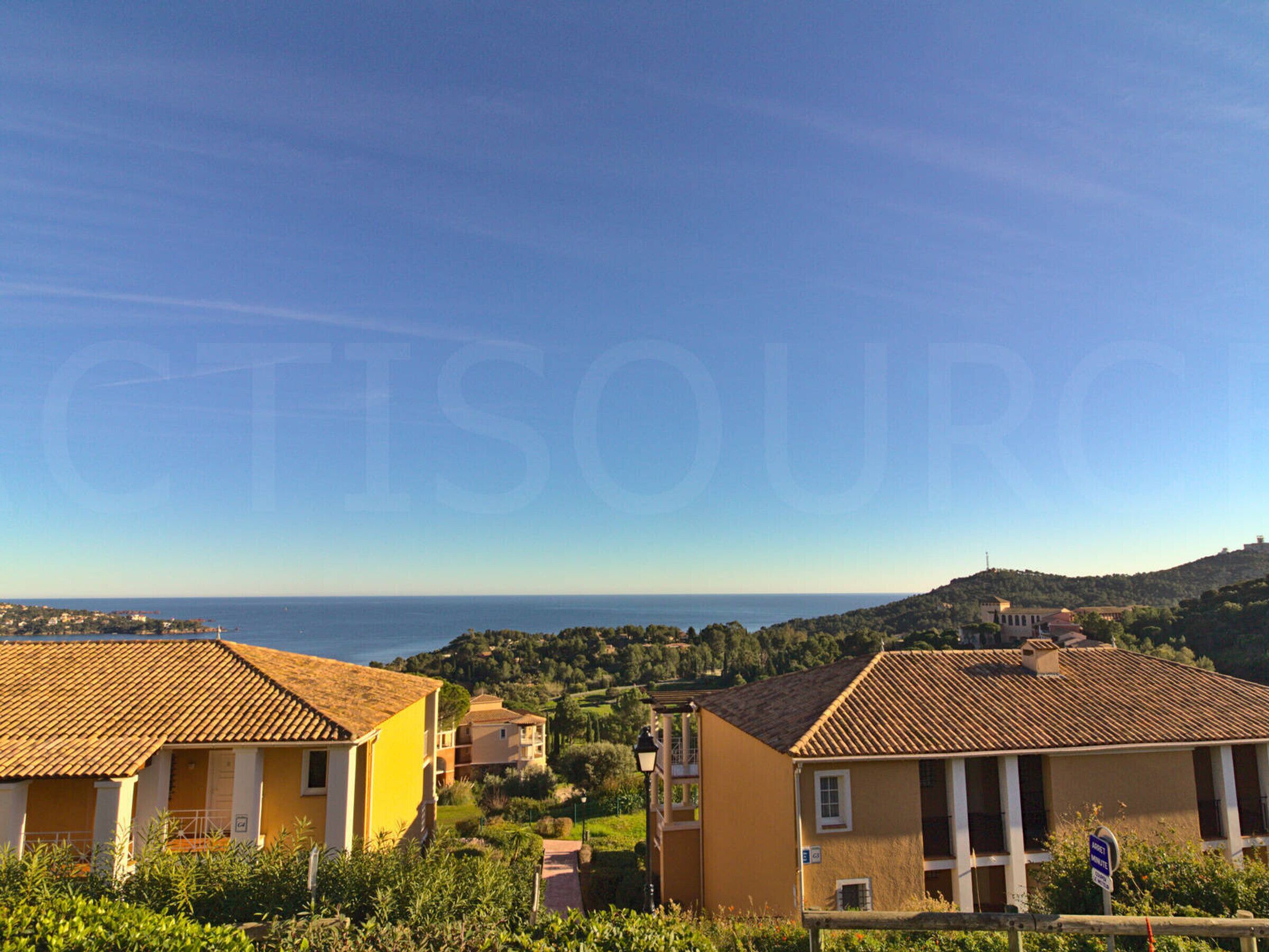 Uma vila costeira com edifícios de telhados em terracota, céu claro e vista para o mar.