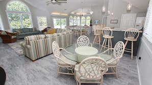 A spacious living room with a large sofa set, a glass coffee table, and wicker chairs. There are multiple windows providing natural light and a view of the outdoors. - Soundfront, Sound View, Hot Tub, Community Amenities, 528, Celestial Haven (Corolla)