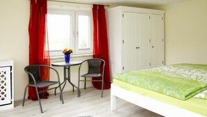 A bedroom with a bed, a window with red curtains, a white wardrobe, and a small table with two chairs.