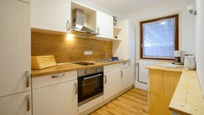 A modern kitchen with white cabinets, a wooden countertop, a built-in oven, and a window with a curtain. - Reiherweg 1, Wohnung EG (Sylt)