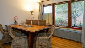 A dining area with a wooden table and wicker chairs, a potted plant, and a window with blinds and a sailboat model. - Reiherweg 1, Wohnung EG (Sylt)
