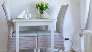 A dining area with a white table, wicker chairs, a wall clock, and a vase with flowers. - Schoenhier-timmendorf (Timmendorfer Strand)