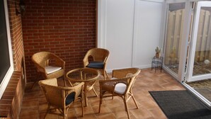 Une terrasse ensoleillée, meublée de meubles en osier et dotée d’une table en verre, avec un mur en briques et une porte-fenêtre coulissante donnant sur l’extérieur.