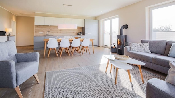 Ein modernes Wohnzimmer mit grauem Sofa, einem Essbereich mit weißen Stühlen, einer Küche mit Holzschränken und einem Kaminofen.
