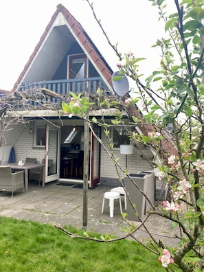 A house with a sloped roof, a satellite dish, a dining area with chairs and a table, and a garden with blooming flowers.