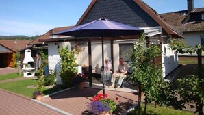 A covered outdoor seating area with a patio table and chairs, surrounded by potted plants and a barbecue grill. - Ferienhaus Pahl in Wolfshagen (Langelsheim)