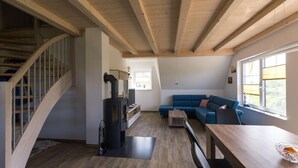 A modern living room with a wooden staircase, a built-in wood stove, a blue sofa, and a dining table. - Fewo Hiddenseeblick (Dranske)
