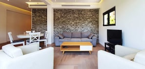 Uma sala de estar moderna com uma parede de destaque em pedra, um sofá cinza, poltronas brancas, uma mesa de centro de madeira e uma televisão de tela plana.