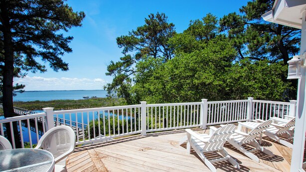 A wooden deck with white railings, a table, and chairs, overlooking a body of water and trees. - Soundfront, Sound views, Private Pool, Hot Tub, MS12, Sounds Amazing (Corolla)