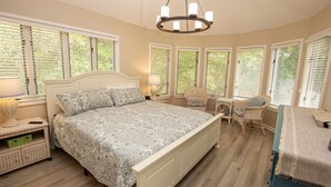 A bedroom with a large bed, multiple windows with blinds, a chandelier, wicker chairs, and a small table with a lamp. - Soundfront, Sound views, Private Pool, Hot Tub, MS12, Sounds Amazing (Corolla)