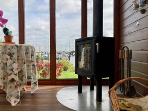 A room with a large window, a wooden table with a floral tablecloth, a black wood stove, and a wicker chair. - 4pers - House w Sauna, Winter Garden & Fishing Pier in Front of the Lauwersmeer (Anjum)
