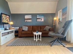 A living room with a brown sofa, a white table, and a grey armchair. There are two wall-mounted pictures, a television, and a lamp.