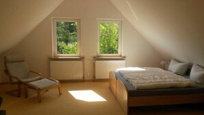 A bedroom with a sloped ceiling, two windows with a view of greenery, a bed with a white duvet, a chair, and a radiator. - Ferienhaus Jana (Wernigerode)