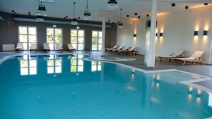 Ein Innenpool mit klarem, blauen Wasser, umgeben von Liegestühlen und Sonnenliegen, mit großen Fenstern für natürliches Licht.