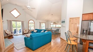 A spacious living room with a blue sofa, wooden flooring, a dining area with chairs, and a kitchen with wooden cabinets and a refrigerator. - Oceanside, Hot Tub, Close to Beach, Sunroom, CP7, Shoulda Been Here Yesterday (Corolla)