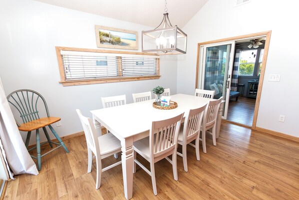 A dining area with a white table and chairs, a chandelier, a window with blinds, and a view of the outdoors.