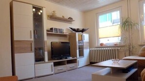 Ein modernes Wohnzimmer mit Fernseher, weißen Schränken, einem Couchtisch und einem Fenster mit einer Pflanze.