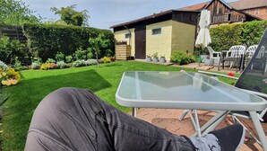 Ein Garten mit Glastisch und -stühlen, einer Hecke und einem Haus im Hintergrund.