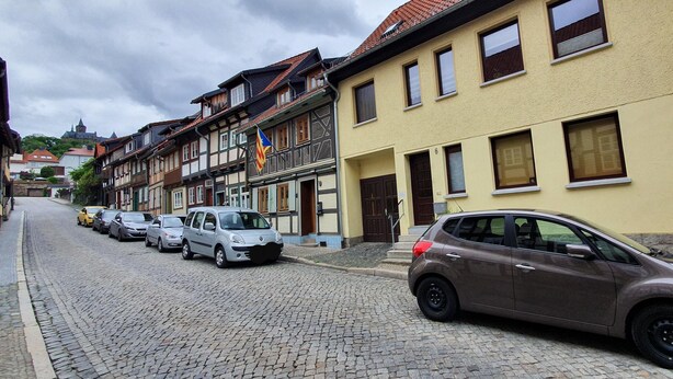 Eine Kopfsteinpflasterstraße gesäumt von traditionellen europäischen Gebäuden, parkenden Autos und einer Flagge.