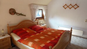 Une chambre à coucher avec un lit en bois, deux tables de chevet, une commode, une fenêtre ornée de rideaux à motifs floraux et un radiateur.