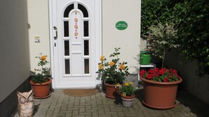 A white door with a glass panel, flanked by potted plants with yellow and red flowers, and a green sign on the wall. - Weißes Haus am Kurpark, Fewo Waldblick (Quedlinburg)