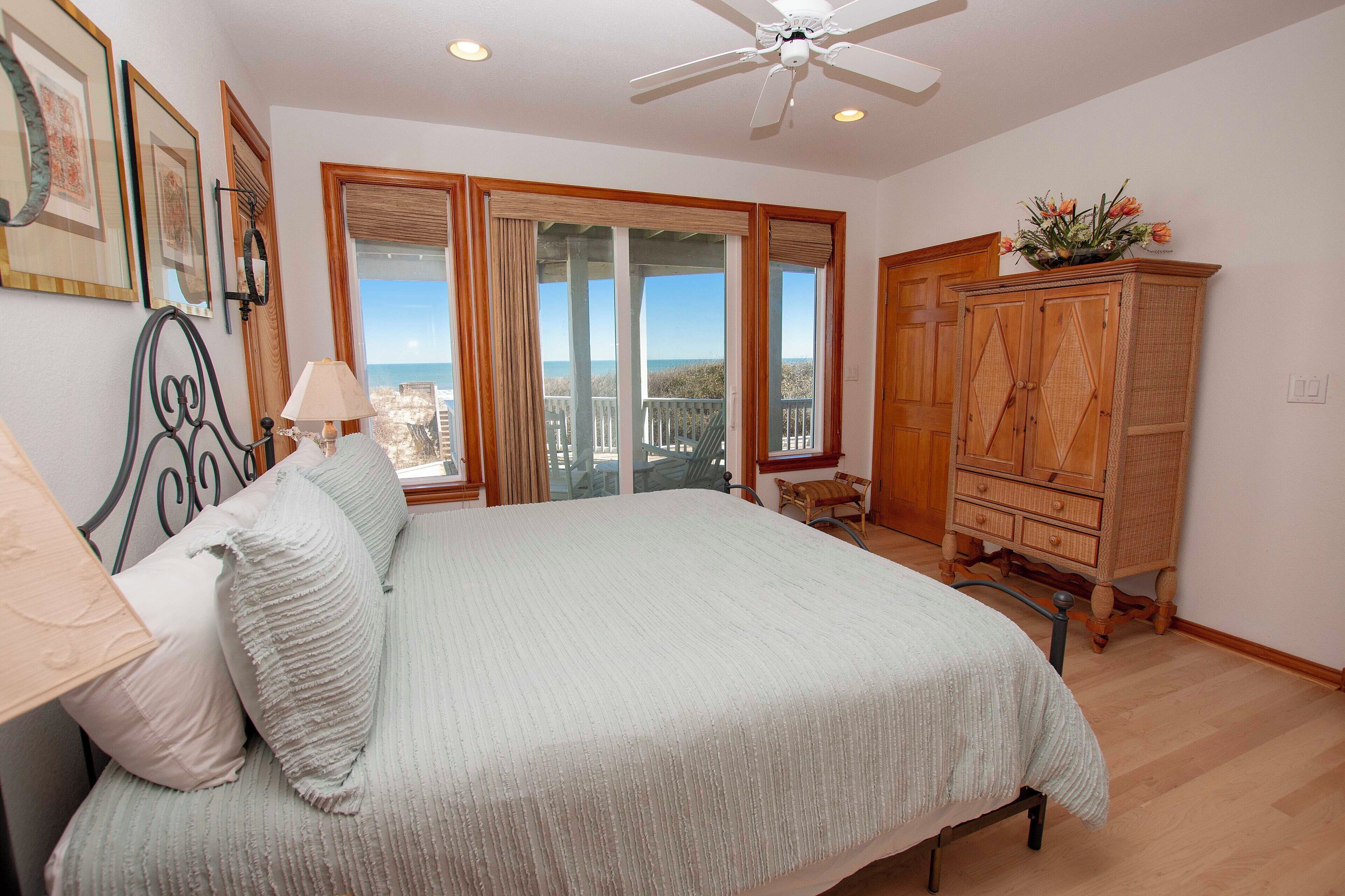 A bedroom with a large bed, a ceiling fan, a wooden cabinet, and a view of the outdoors through a sliding door.