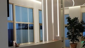A reception desk with a modern design, featuring a curved countertop and a blue wave pattern at the base. There are large windows with blinds, and a potted plant is visible in the background.