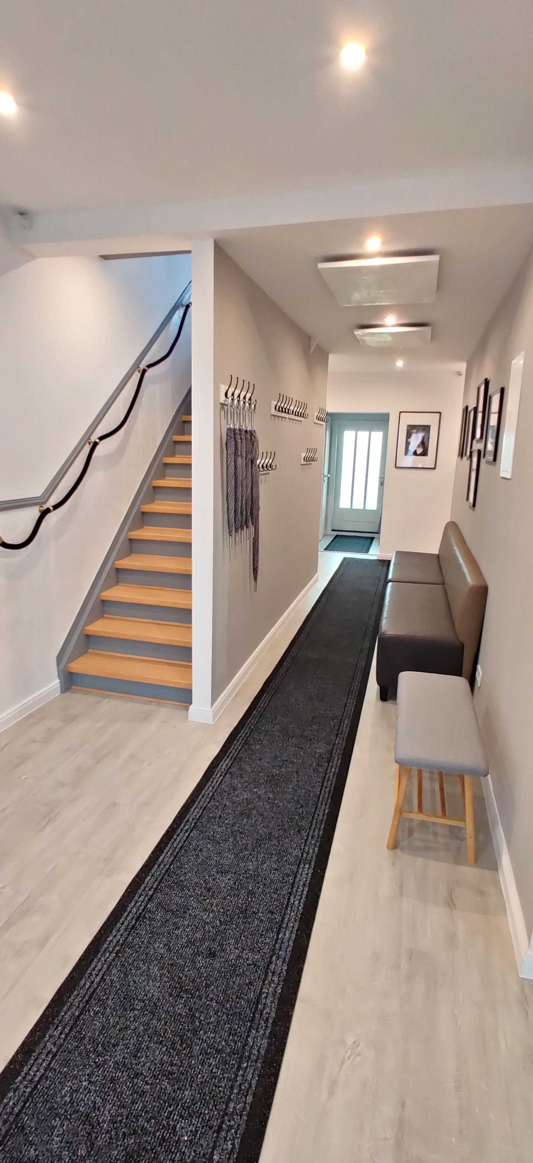 A modern interior with a staircase, a hallway leading to a door, a bench, and a wall-mounted coat rack.