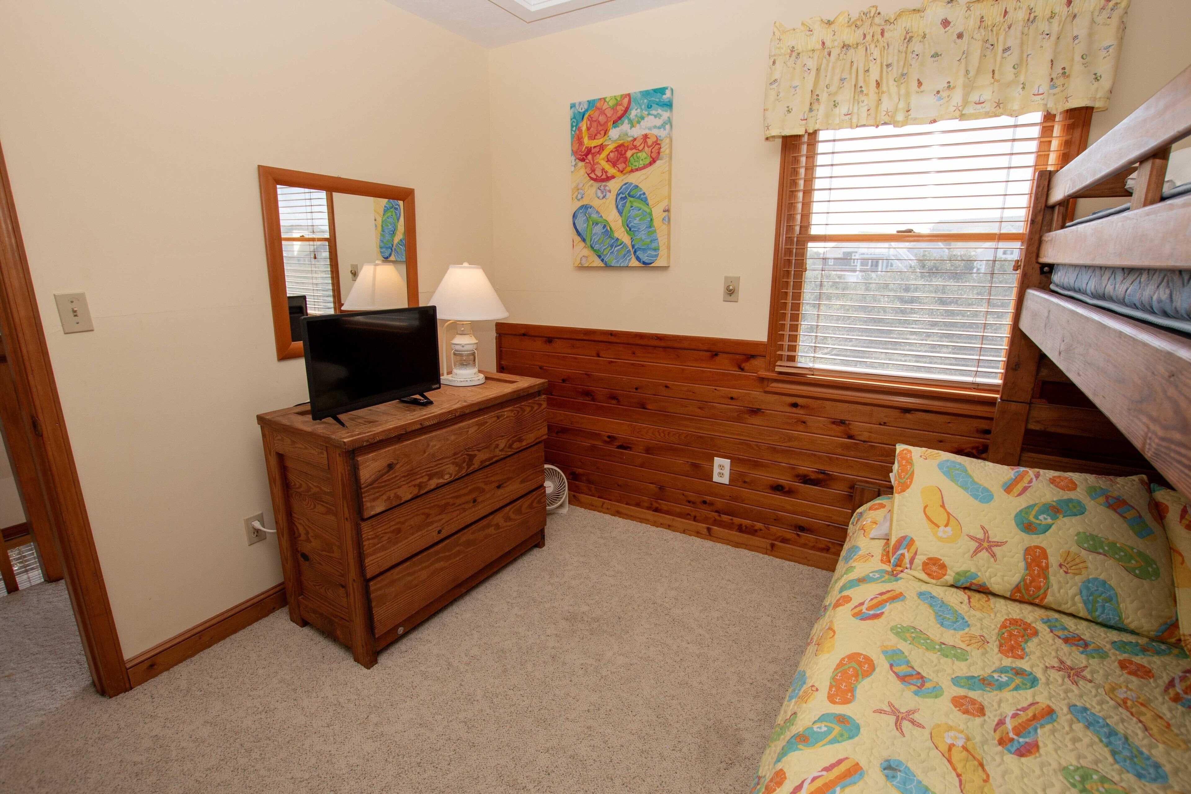A bedroom with a wooden dresser, a bed with a patterned bedspread, a window with blinds, a wall-mounted TV, and a mirror.