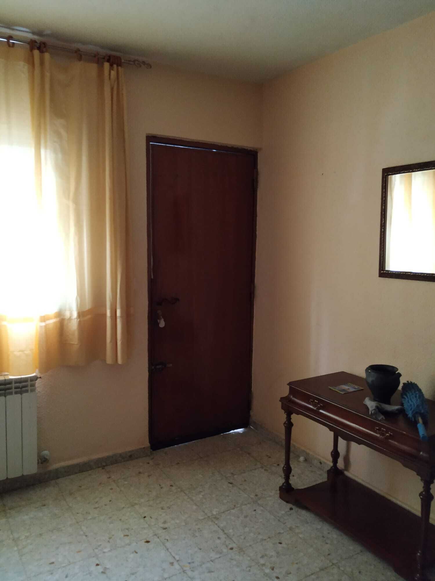 A room with a closed wooden door, a radiator, a small table with a vase and a brush, and a window with curtains.