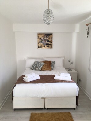 A neatly made bed with white linens, a brown throw blanket, and two pillows. There is a small bedside table with a decorative item on it. - Ashdown Apartment (Corby)