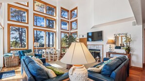 A spacious living room with a large window wall, a fireplace, and a television. - Soundfront, Amazing Sound Views! Sunsets, Yard, Hot Tub, MS100, Timberly II (Corolla)