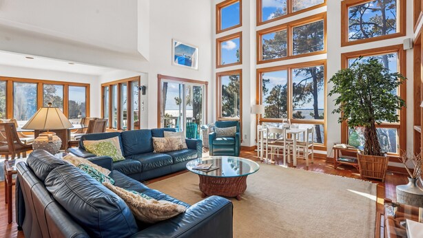 A spacious living room with a large blue sofa, a glass coffee table, and multiple windows offering views of trees and the sky. - Soundfront, Amazing Sound Views! Sunsets, Yard, Hot Tub, MS100, Timberly II (Corolla)