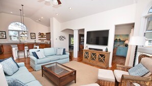A spacious living room with a light blue sofa, a wooden coffee table, and wicker furniture. A large flat-screen TV is mounted on a wooden cabinet.