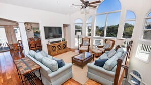 A spacious living room with a large TV, comfortable seating, and a balcony with a view.