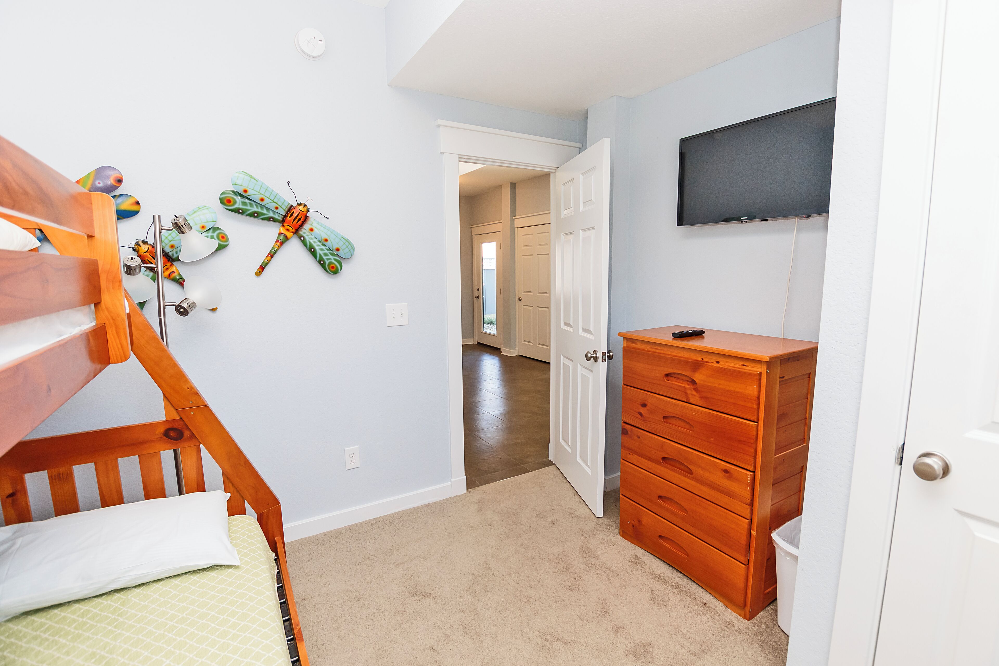 A bedroom with a bunk bed, a wooden dresser, a TV mounted on the wall, and a door leading to another room.