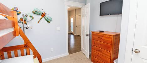 A bedroom with a bunk bed, a wooden dresser, a TV mounted on the wall, and a door leading to another room.