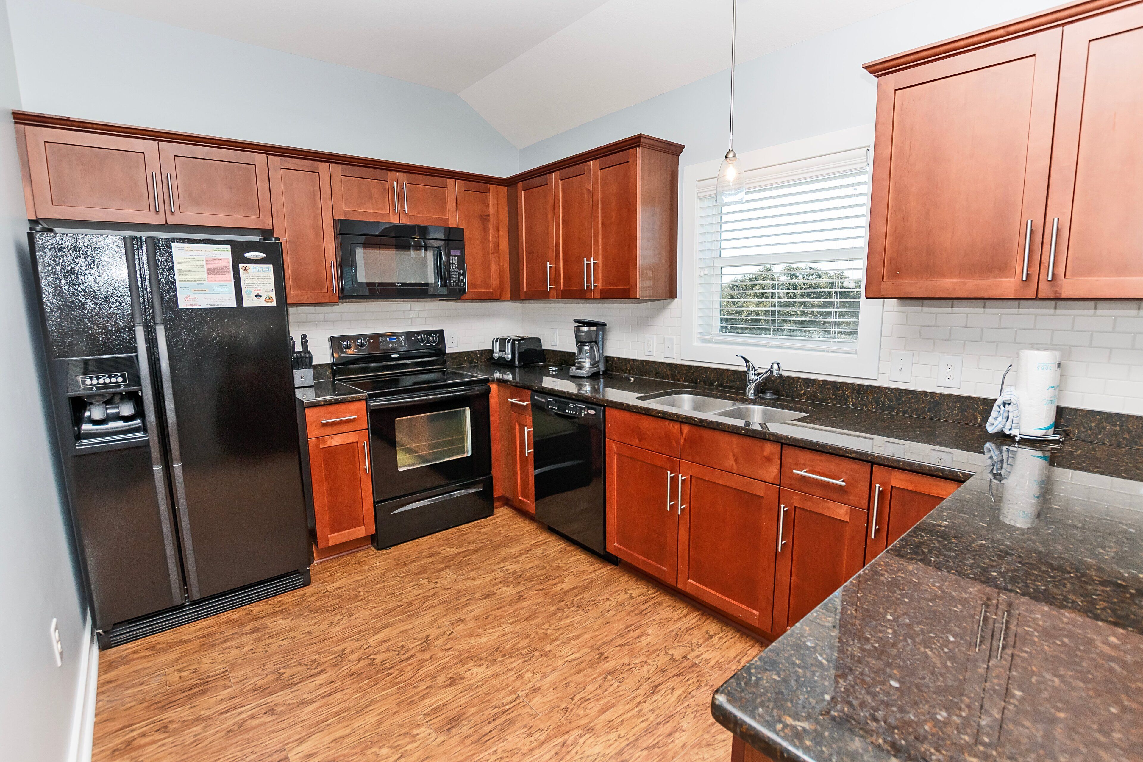 A modern kitchen with wooden cabinets, a black refrigerator, a microwave, a dishwasher, and a granite countertop.