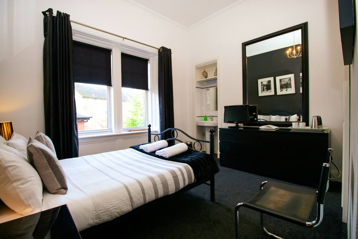Quarto com cama, escrivaninha, cadeira, espelho e janela com cortinas.