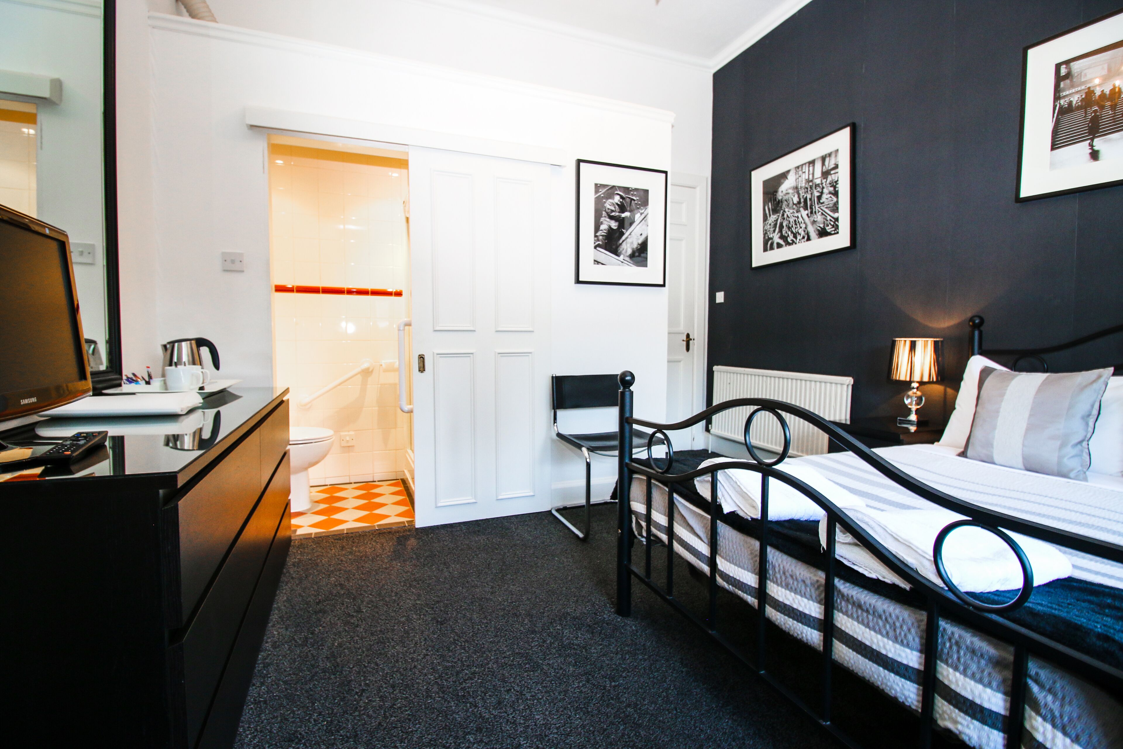 A hotel room with a bed, a desk, a chair, a radiator, and a door leading to a bathroom.