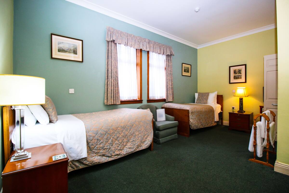 Quarto de hotel com duas camas, uma escrivaninha, uma cadeira, um abajur e um cabideiro.