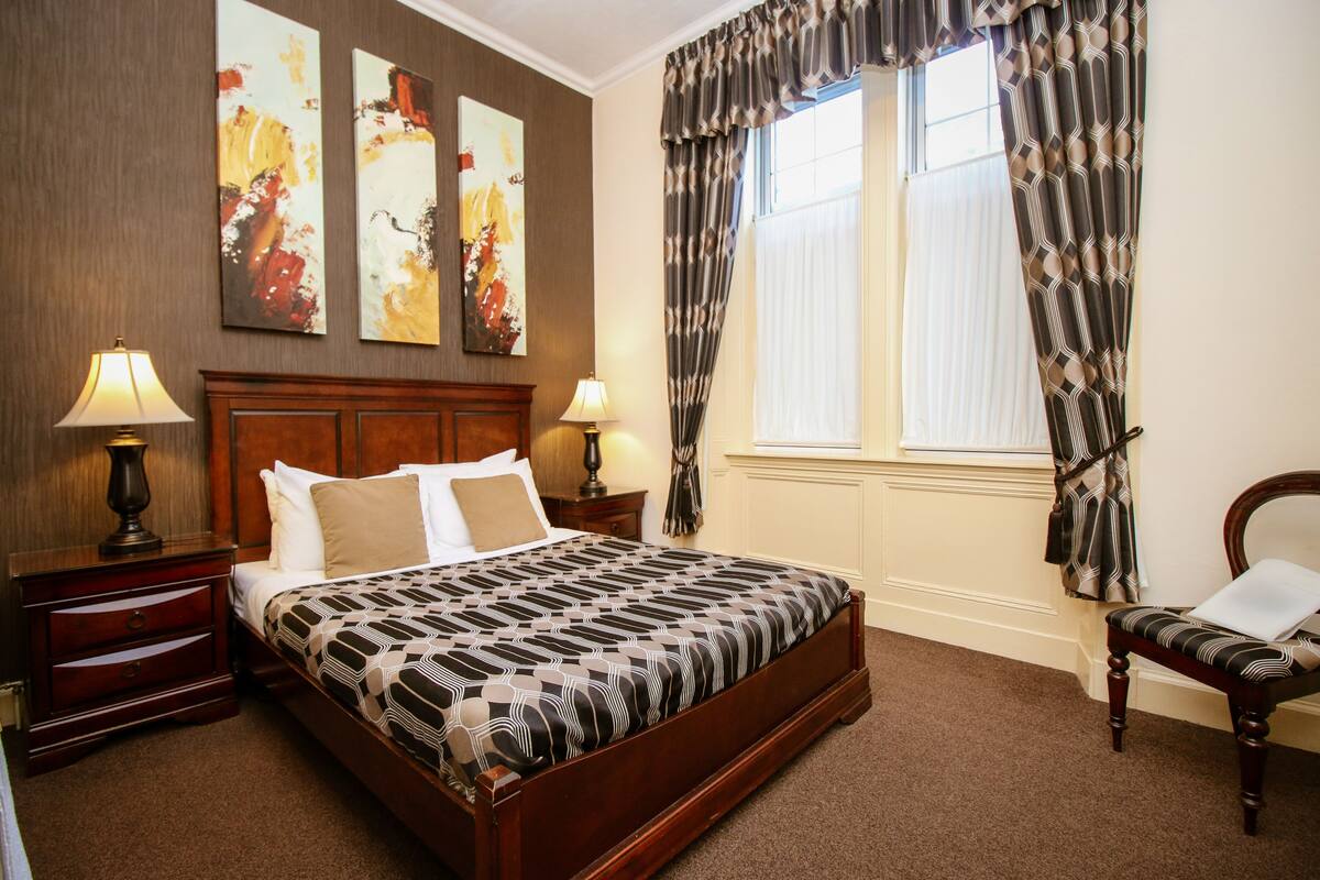 Quarto de hotel bem arrumado com uma cama grande, duas mesas de cabeceira com abajures, uma poltrona e uma janela com cortinas.