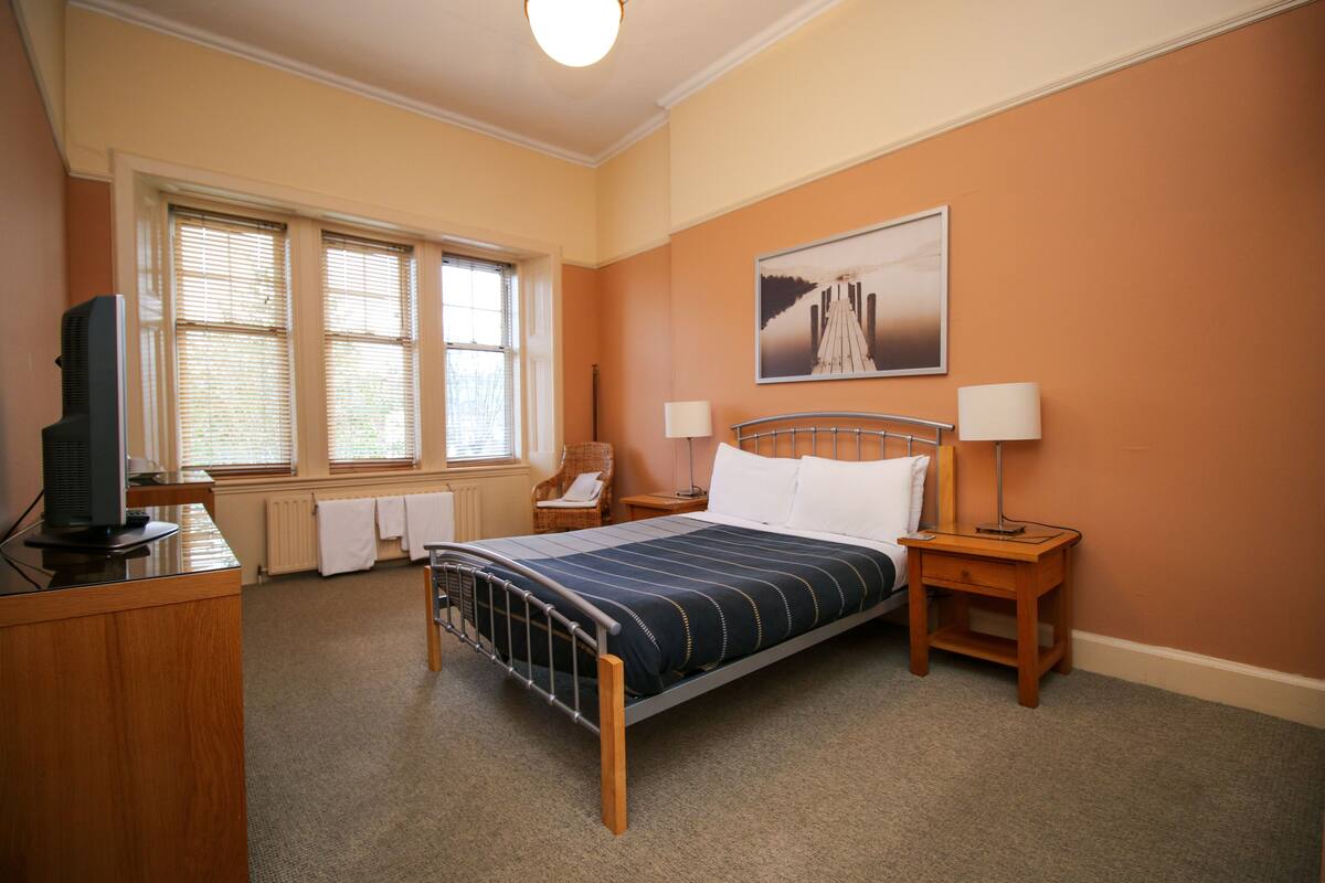 Quarto de hotel com uma cama, duas mesas de cabeceira com luminárias, uma cadeira, uma escrivaninha com televisão e uma janela com persianas.