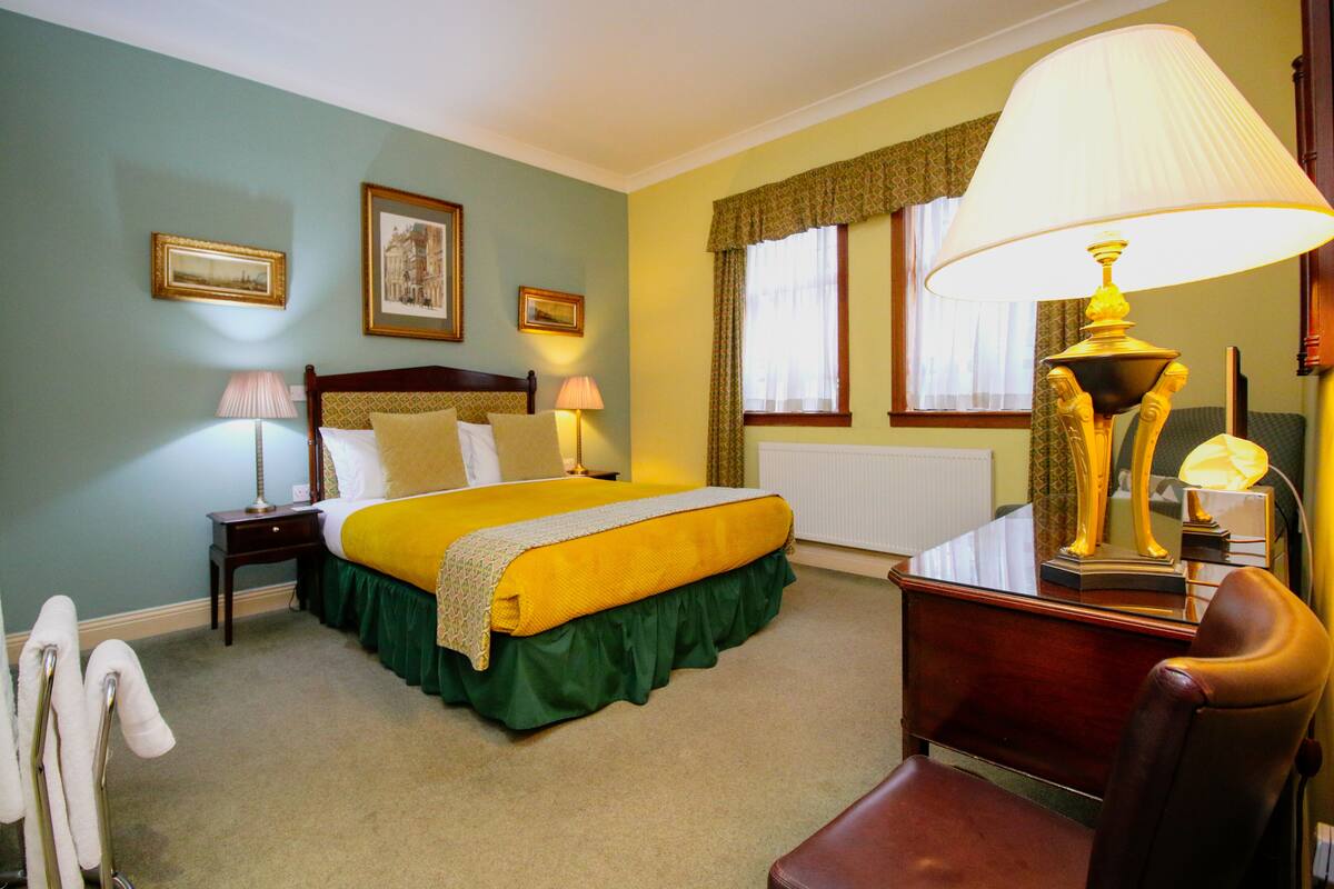 Quarto de hotel com uma cama, duas luminárias de cabeceira, uma escrivaninha com abajur, uma cadeira e um radiador.